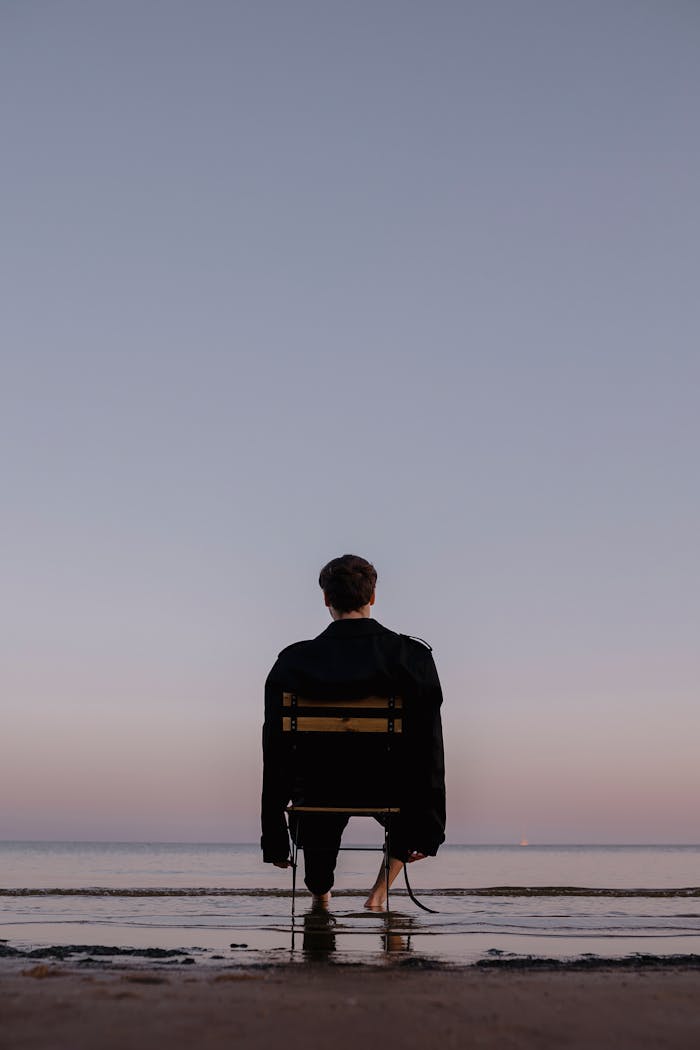A man sits on a chair facing the calm sea at dusk, creating a tranquil moment.